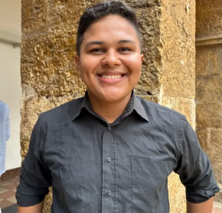 Deivid, discípulo na Comunidade Maíra vestindo uma camisa escura sorri enquanto está de pé, tendo como fundo uma parede de pedra.