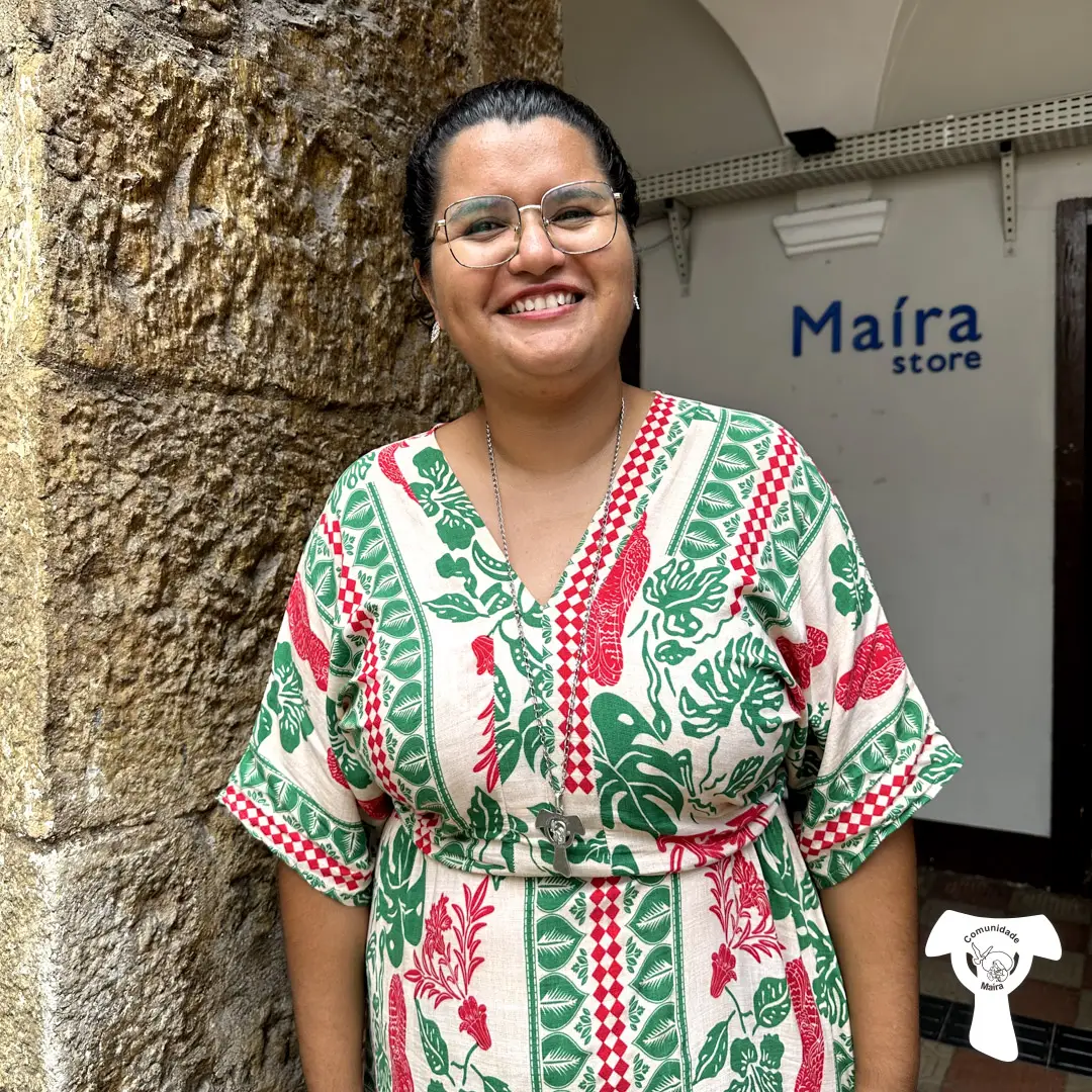 Letícia, consagrada de Vida, sorrindo, vestindo um vestido verde e vermelho com estampa florida, transmitindo alegria. Ao fundo uma parede rústica de pedra.