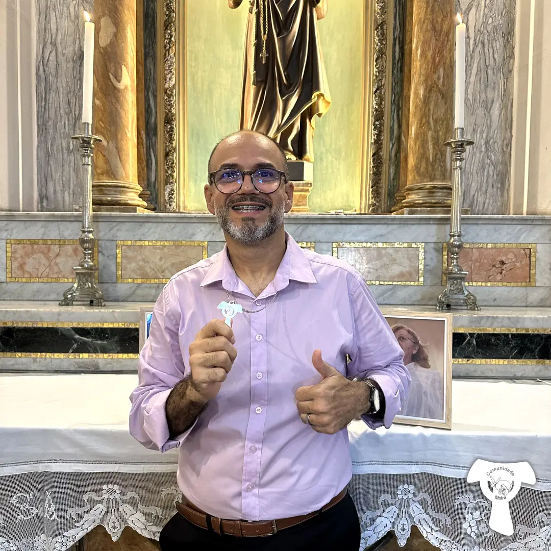 Wagner, consagrado de Aliança, vestindo camisa social rosa está em frente a um altar segurando o Tau da Comunidade e fazendo sinal de positivo.